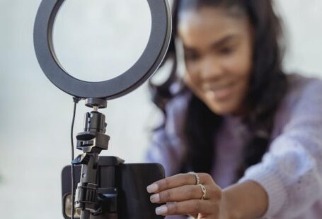Tutorials - Cheerful young African American female blogger in stylish sweater smiling while setting up camera of smartphone attached to tripod with ring light before recording vlog
